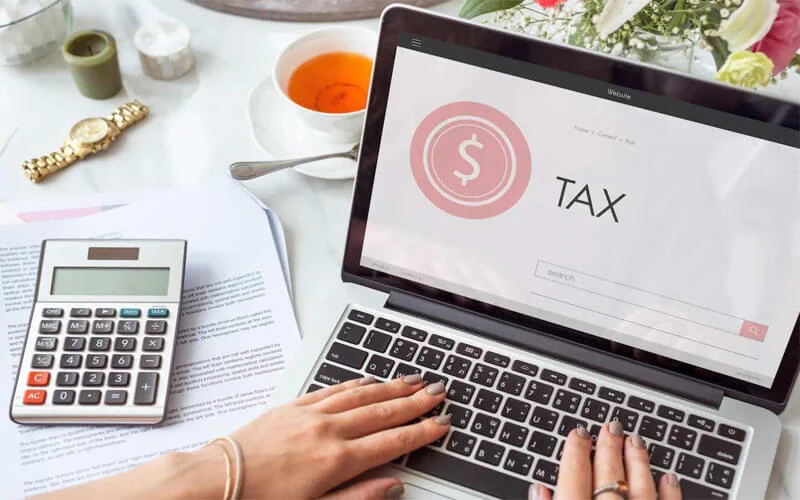 Người thao tác trên máy tính xách tay hiển thị chữ TAX, bên cạnh máy tính cầm tay và giấy tờ.